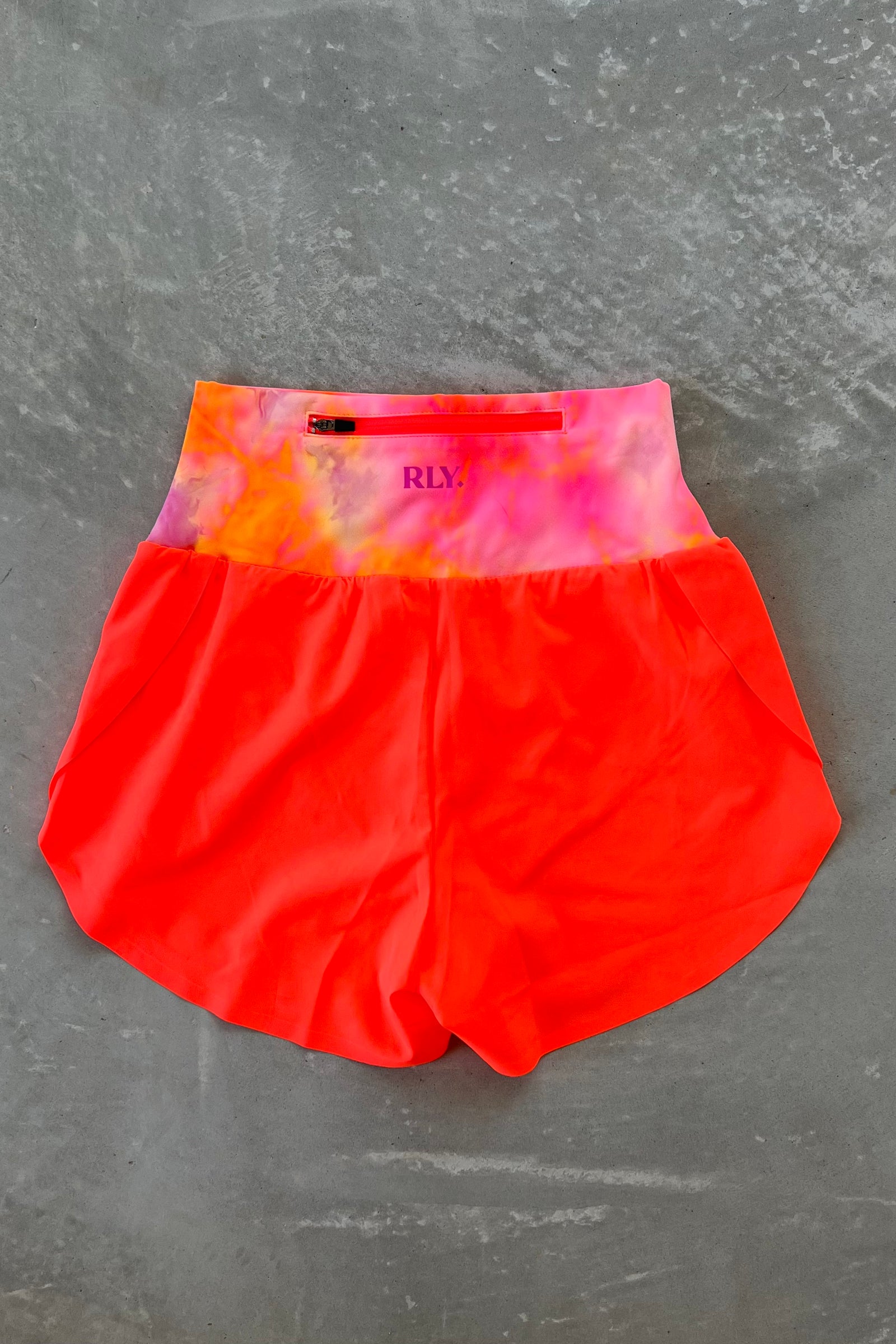 A pair of tie-dye split shorts with the top panel featuring a sunburst pattern and the bottom panel in neon coral color. The shorts are designed for active wear and have a high waist.