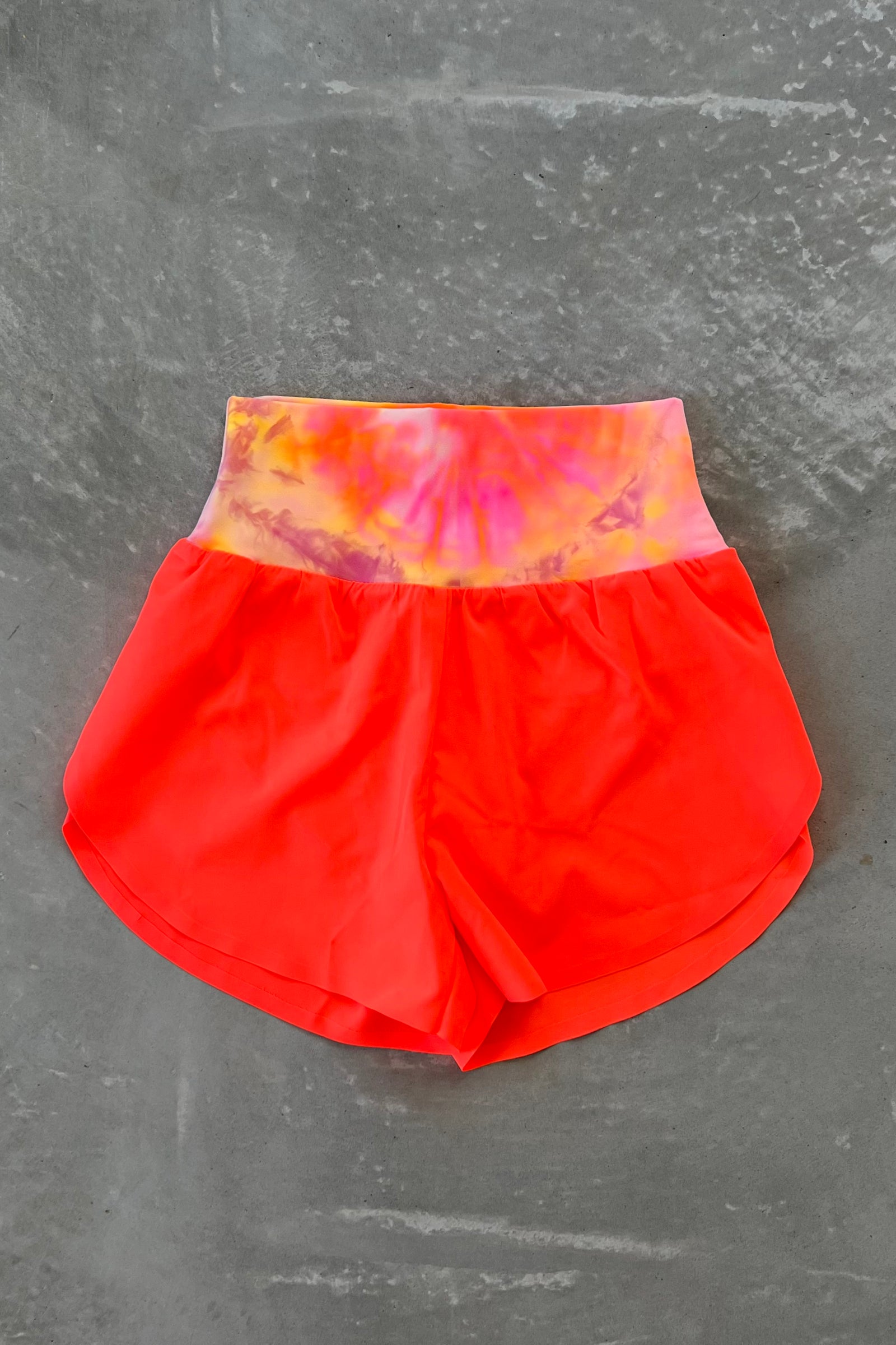 A pair of tie-dye split shorts with the top panel featuring a sunburst pattern and the bottom panel in neon coral color. The shorts are designed for active wear and have a high waist.